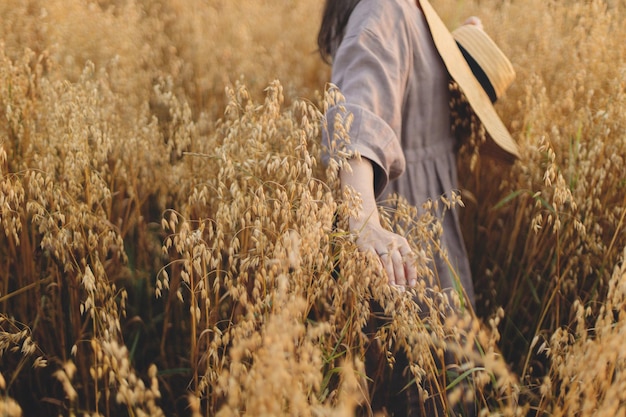 Stylish woman with straw hat holding oat stems in evening light Rural slow life Young female in rustic linen dress standing in harvest field in summer countryside Atmospheric tranquil moment