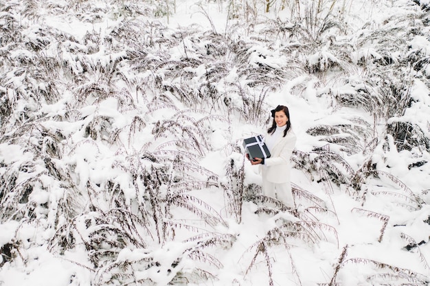 A stylish woman with a white suit with a New Year's gift in her hands in a winter forest A girl in nature in a snowy forest with a gift box