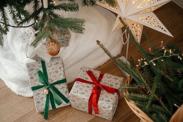 Stylish wrapped christmas gifts and rustic basket with fir branches under festive decorated christmas tree in scandinavian room Merry Christmas and Happy holidays