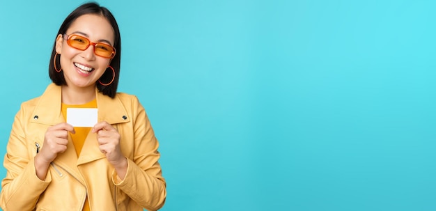 Stylish young asian woman in sunglasses showing credit card and smiling recommending bank contactless payment or discounts in store standing over blue background