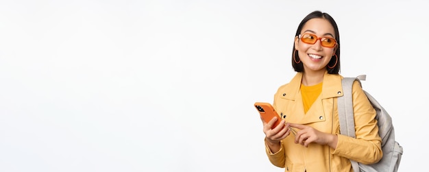 Stylish young asian woman tourist traveller with backpack and smartphone smiling at camera posing against white background