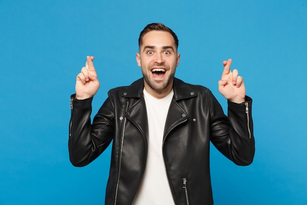 Stylish young bearded man in black leather jacket white t-shirt waiting for special moment, keeping fingers crossed isolated on blue wall background. People sincere emotions lifestyle concept. Mock up