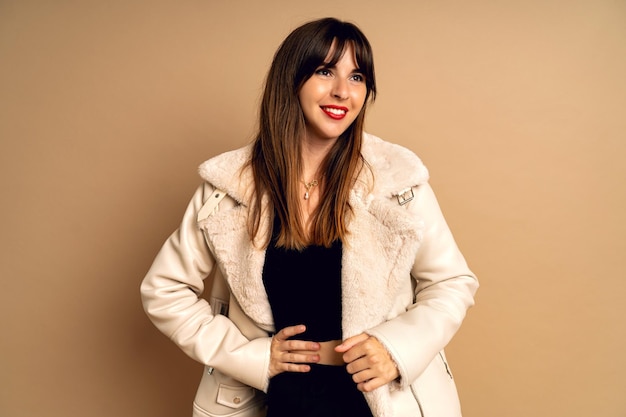 Stylish young brunette woman posing at fur coat beige studio background winter time