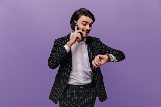 Stylish young brunettehaired businessman in light shirt and striped suit considering time by phone looking at watch Busy man posing isolated on violet background