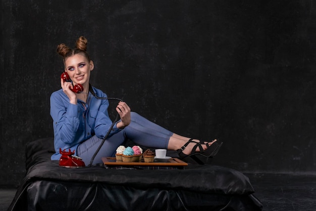 Stylish young businesswoman sits on bed with phone in her hands and dines on cupcakes. Black background