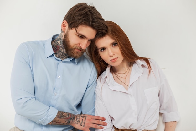 Stylish young couple beautiful trendy redhead woman and handsome hipster brutal business man with beard and tattoos stand near white background