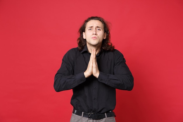 stylish young curly long haired man in black shirt posing isolated on red wall background studio portrait. People sincere emotions lifestyle concept. Mock up copy space. Hands folded in prayer gesture