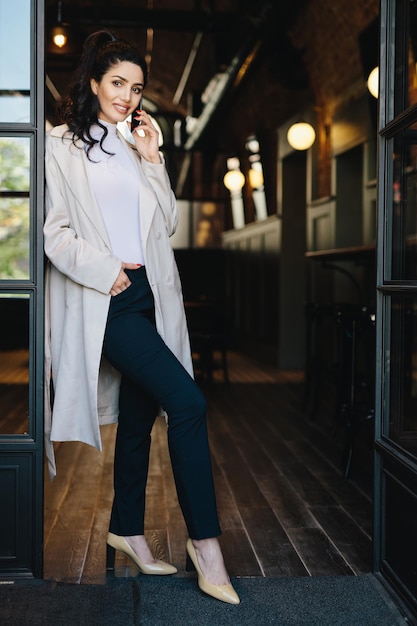 Stylish young female with dark hair wearing white coat black trousers and shoes with heels posing into camera while standing against cafe background chatting over phone holding hand in pocket