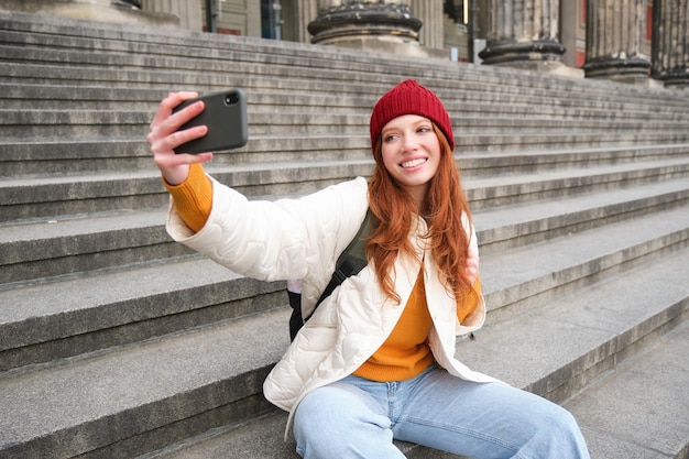 Stylish young girl in red hat takes photos on smartphone camera makes selfie as she sits on stairs n