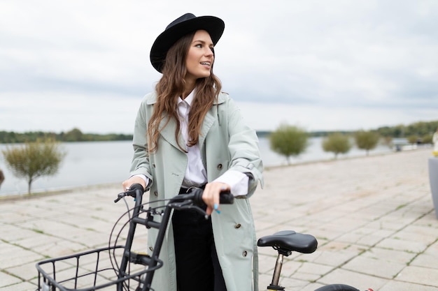 Stylish young girl with a bicycle in a black hat on the embankment