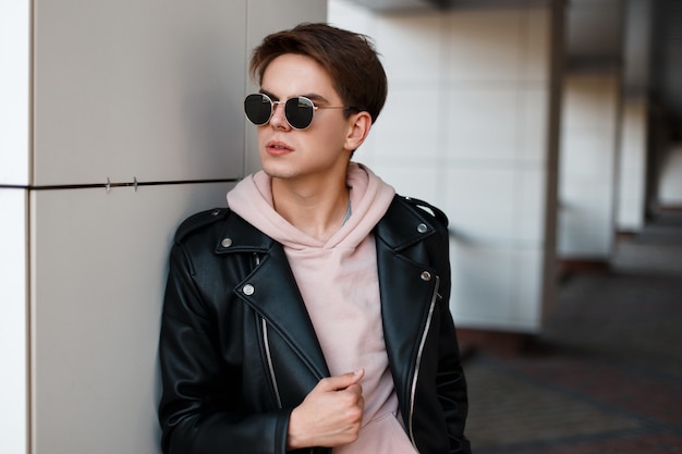 Stylish young hipster man with a fashionable hairstyle in stylish black glasses in a black leather jacket in a pink sweatshirt stands near a white wall. Attractive american guy. Men's fashion.