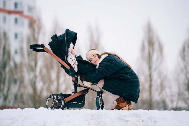 stylish young mother hugs her daughter sitting in stroller in park outdoor at winter.