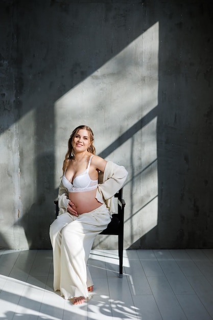 A stylish young pregnant woman in a white suit is sitting on a chair on a gray background Pregnant woman in white underwear