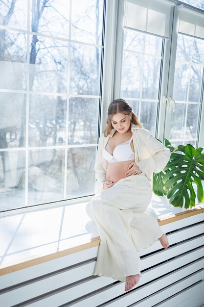 A stylish young pregnant woman in a white suit is sitting on the windowsill Pregnant woman in white underwear