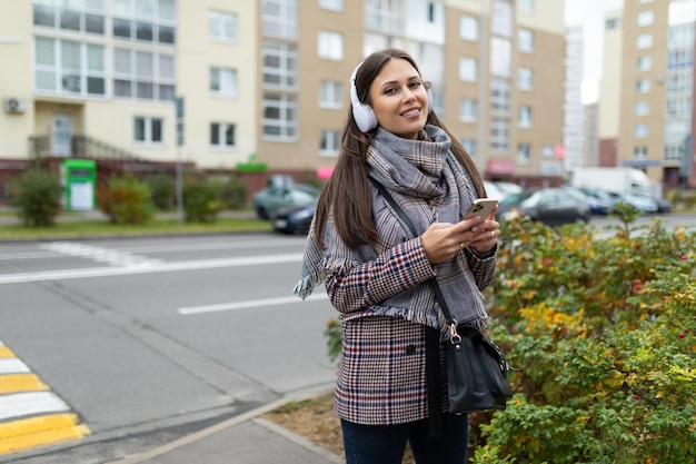 Stylish young woman in an autumn coat walks around the city listening to music in headphones with a