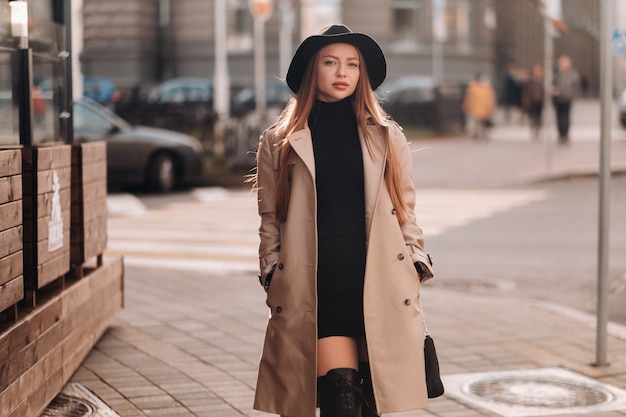 Stylish young woman in a beige coat in a black hat on a city street. Women's street fashion. Autumn clothing.Urban style