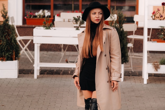 Stylish young woman in a beige coat in a black hat on a city street. Women's street fashion. Autumn clothing.Urban style.