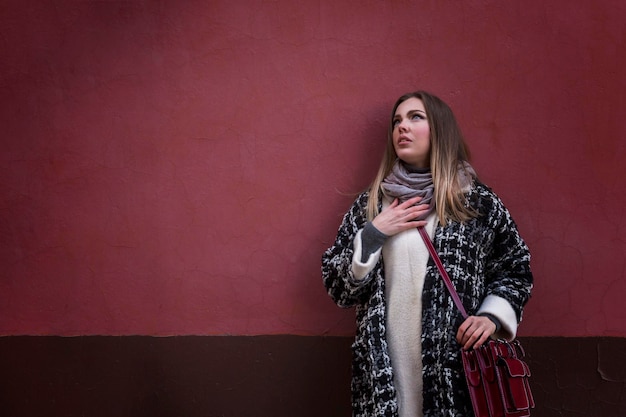 A stylish young woman in a coat looks up in surprise on the street in the city against the backdrop of a red brick building Space for text