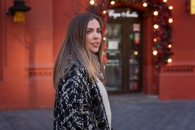 A stylish young woman in a coat walks in a city decorated for Christmas New Year holidays Beautiful shop windows and cafes in a red brick building Closeup