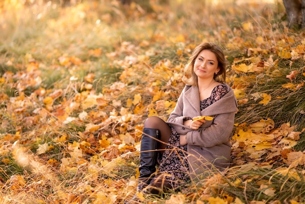 Stylish young woman in a gray coat sits on yellow leaves in an autumn park