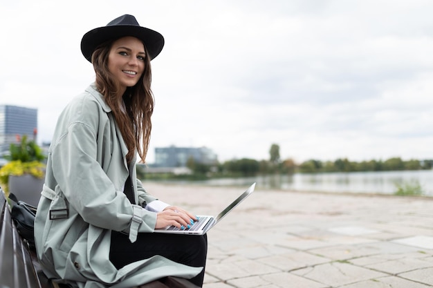 Stylish young woman in hat working on laptop outside