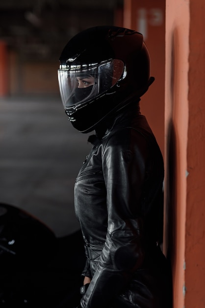 Stylish young woman motorcycle rider in black protective gear and full-face helmet near her bike