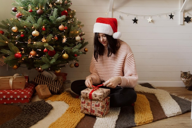 Stylish young woman opening christmas gift box with cat under christmas tree with lights in modern room Happy girl in santa hat holding christmas presents Happy holidays