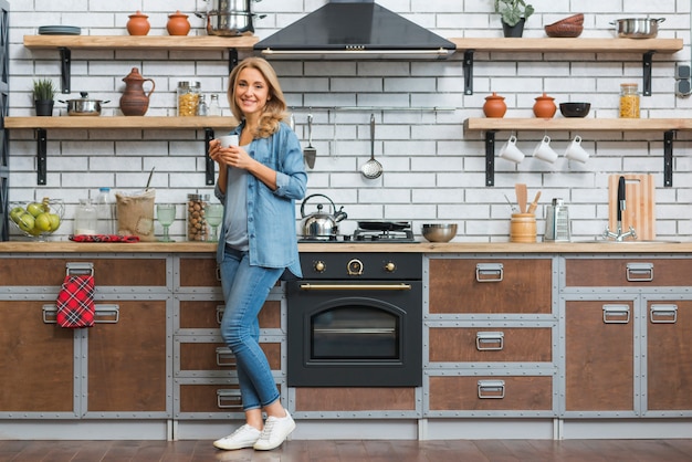 Photo stylish young woman standing in modular kitchen holding cup of coffee in hand