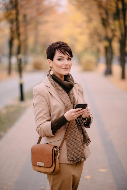 Stylish young woman uses smartphone against background of autumn street