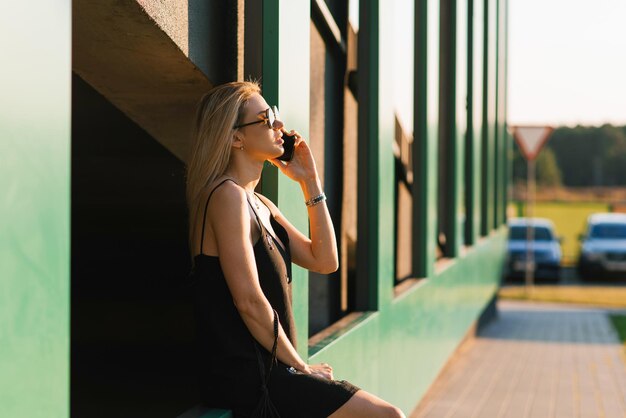 Stylish young woman walking around the city is resting sitting on the parking wall and talking on her smartphone