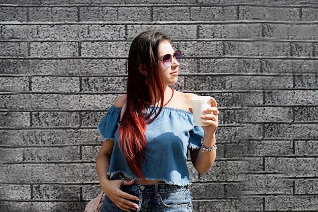 Stylish young woman wearing jeans shirt sunglasses and bag drinking coffee to go at street black brick wall background