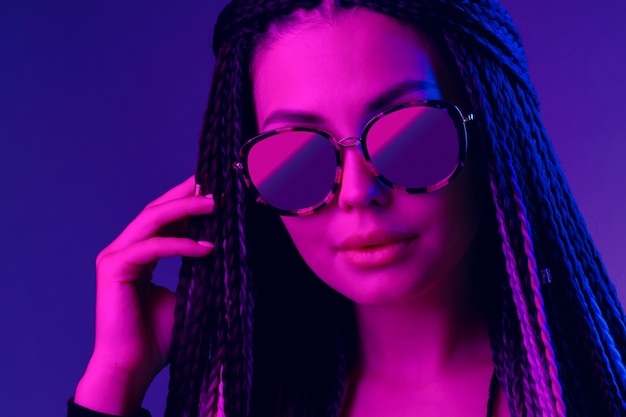 Stylish young woman with braids wearing sunglasses against purple background