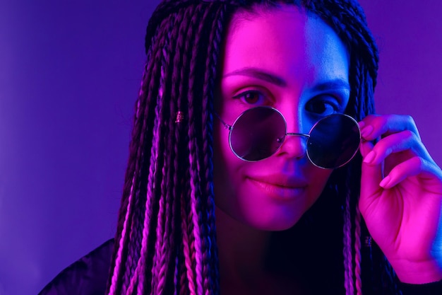 Stylish young woman with braids wearing sunglasses against purple background