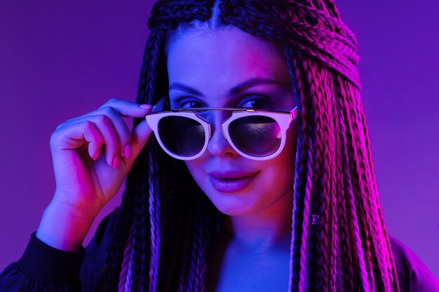 Stylish young woman with braids wearing sunglasses against purple background