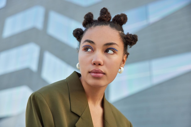 Stylish young woman with hair buns concentrated away with serious expression wears vivid blue eyeliner dressed in green stylish jacket poses against blurred background. Street lifestyle concept