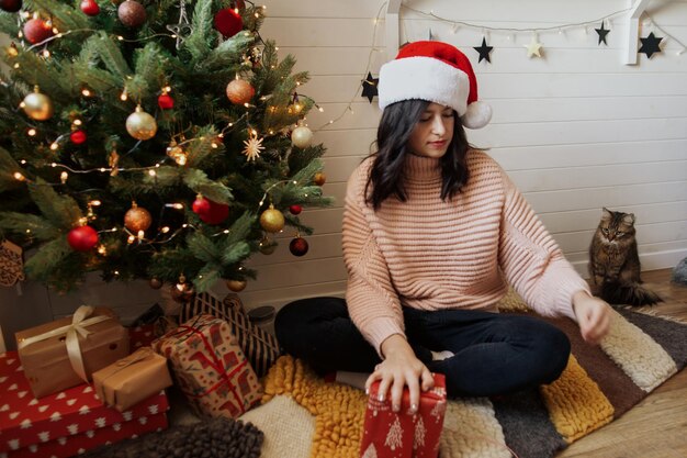 Stylish young woman wrapping christmas present in paper under christmas tree with lights in modern room Happy girl in santa hat and sweater preparing christmas gift