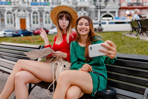 Stylish young women travelling together in Europe dressed in spring trendy dresses and accessories holding map