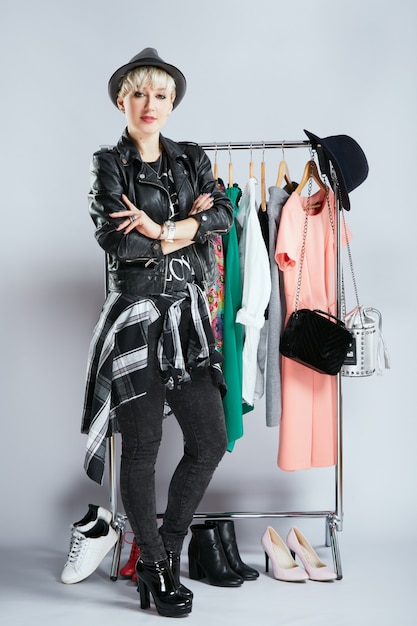 Stylist in fashionable outfit standing near dresses on rack, full body. Person in sphere of fashion choosing clothes, looking at camera. Shopping, indoors, buying of clothes