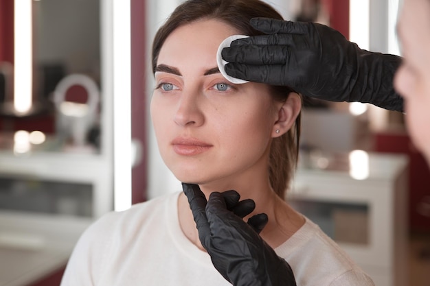 Stylists hands in black gloves wipe the face of the model with a cotton pad Styling of eyebrows