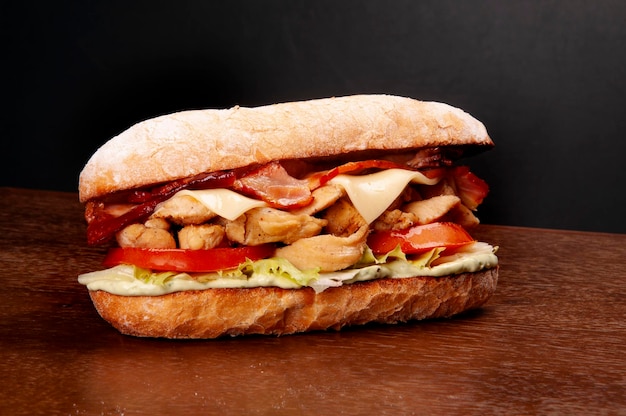 Sub sandwich of chicken cubes bacon lettuce tomato on baguette on wooden table and dark background