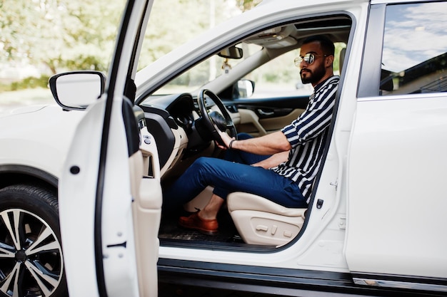 Successful arab man wear in striped shirt and sunglasses pose behind the wheel of  his white suv car. Stylish arabian men in transport.