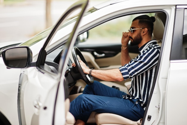 Successful arab man wear in striped shirt and sunglasses pose behind the wheel of  his white suv car. Stylish arabian men in transport.