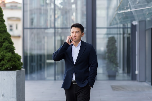 Successful Asian businessman businessman talking on a mobile phone while walking near the office of a large modern, serious and pensive employee