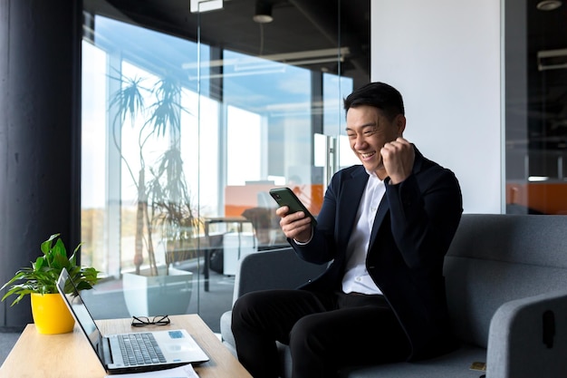 Successful Asian businessman celebrates victory looks at phone happy and rejoices sitting in modern office