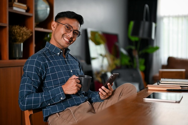 Successful Asian businessman sipping coffee and using his smartphone at his desk