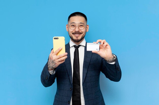 successful asian businessman in suit and glasses holds smartphone and bank card