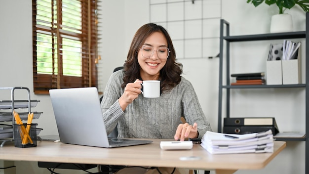 Successful Asian businesswoman or accountant using calculator while sipping coffee at desk