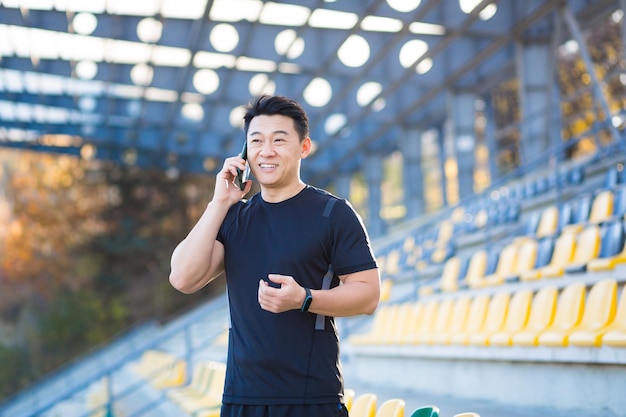 Successful Asian male athlete talking on the phone sharing his athletic achievements and results smiling and rejoicing outside at the sports stadium