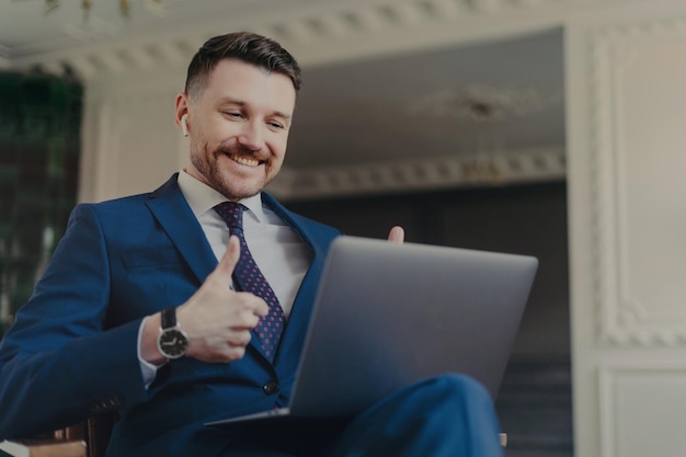 Successful bearded male entrepreneur dressed in formal suit uses modern laptop computer and earbuds for making web conference chatting keeps thumbs raised makes like gesture wears blue formal suit