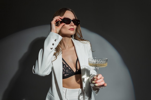 Successful beautiful sexy woman in fashion white suit with blazer and lace bra holds champagne glass and puts on sunglasses on dark background in studio Pretty chic lady celebration event